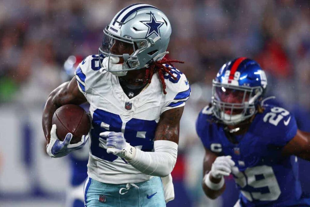 Dynamic Dallas Cowboys player running with football during game, wearing team jersey and helmet, on the field with quarterback and sports fans in background.