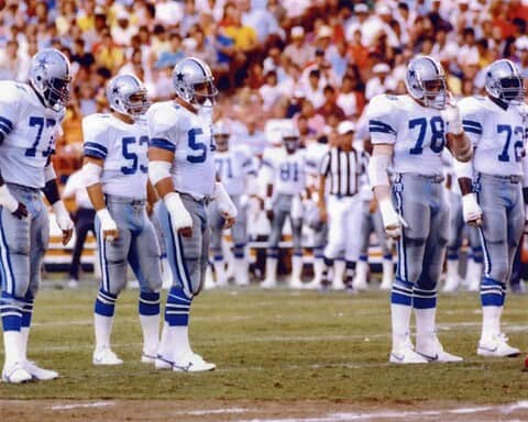 Dallas Cowboys football team standing in formation on the field during a game, wearing white and blue uniforms, with a packed stadium crowd in the background.