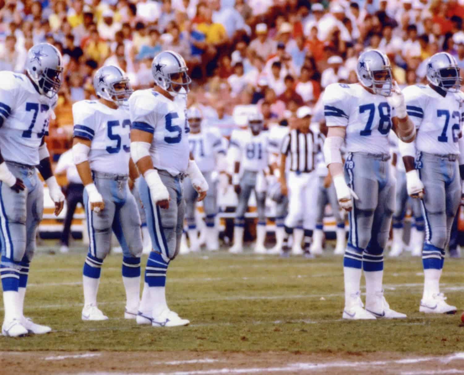 Dallas Cowboys football team standing in formation on the field during a game, wearing white and blue uniforms, with a packed stadium crowd in the background.