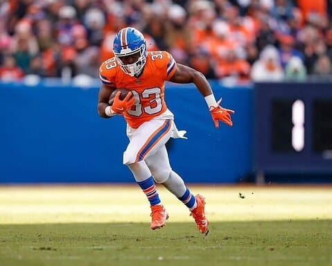 Running back player in orange jersey with football on field, NFL football game, Dallas Cowboys player, football action, stadium crowd in background.