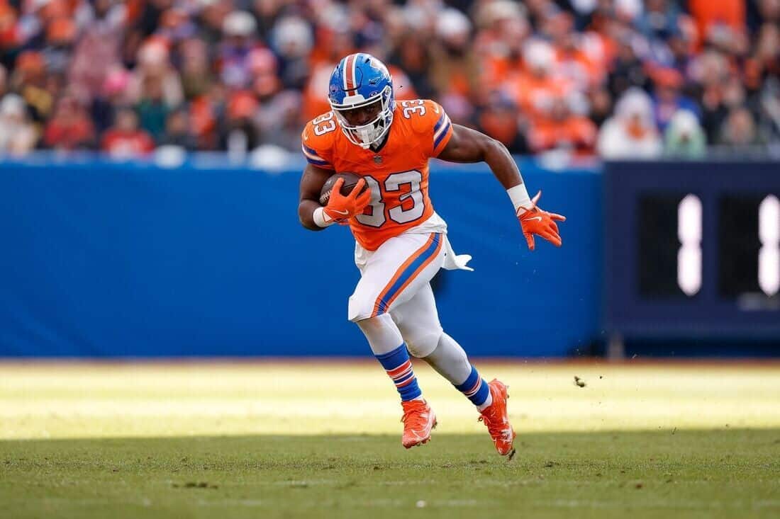 Running back player in orange jersey with football on field, NFL football game, Dallas Cowboys player, football action, stadium crowd in background.