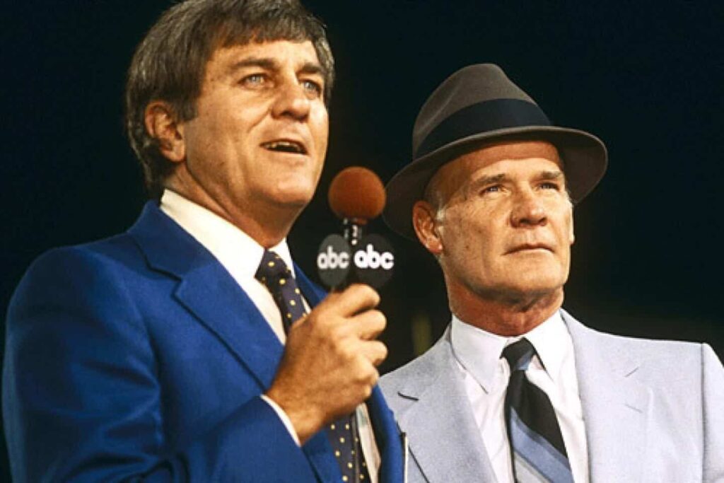 Vince Vaughn and an unidentified man in a gray suit during a Dallas Cowboys broadcast, both dressed in vintage suits and hats, discussing football, inside the Dallas Cowboys stadium.