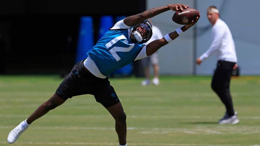 High school football player making a diving catch during practice, wearing a blue jersey with the number 12, on a sunny day at the Dallas Cowboys training facility.