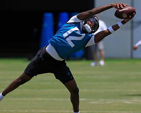 High school football player making a diving catch during practice, wearing a blue jersey with the number 12, on a sunny day at the Dallas Cowboys training facility.