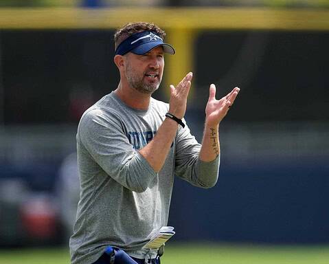 Dallas Cowboys coach speaking on the field during training camp, wearing a grey Cowboys shirt and a blue visor, demonstrating leadership and team strategy.