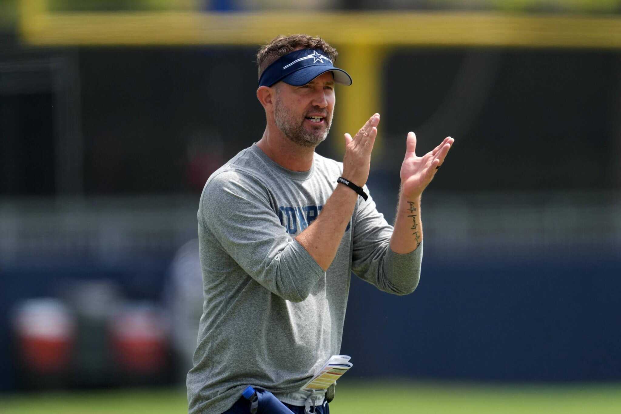Dallas Cowboys coach speaking on the field during training camp, wearing a grey Cowboys shirt and a blue visor, demonstrating leadership and team strategy.