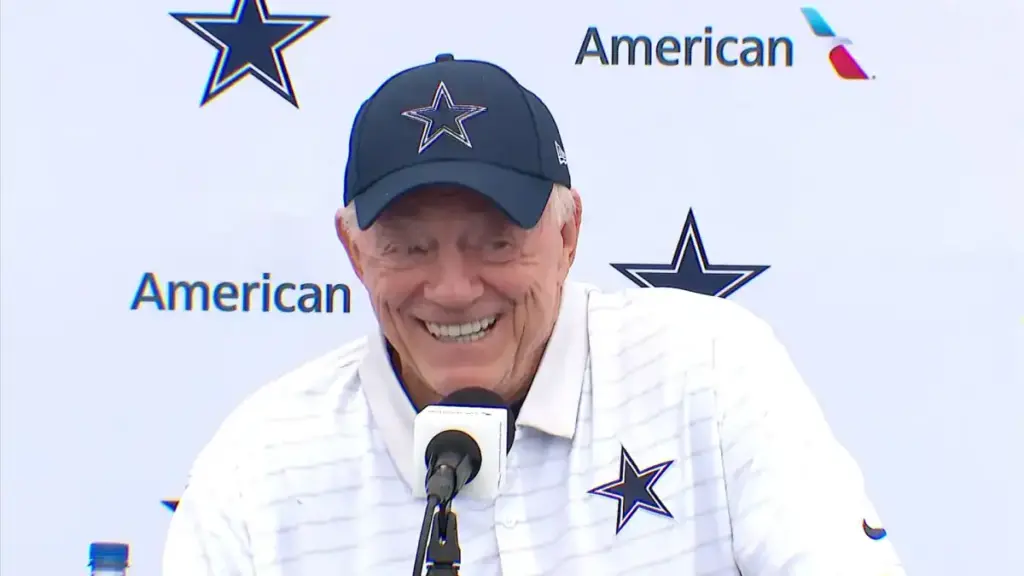 Happy Dallas Cowboys coach Jerry Jones smiling at a press conference during the NFL draft, wearing team apparel with a Cowboys logo, showcasing sports leadership and NFL team branding.