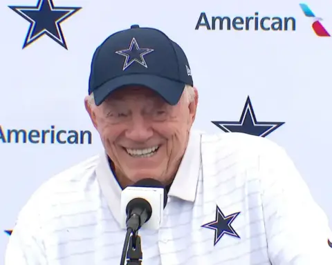 Happy Dallas Cowboys coach Jerry Jones smiling at a press conference during the NFL draft, wearing team apparel with a Cowboys logo, showcasing sports leadership and NFL team branding.