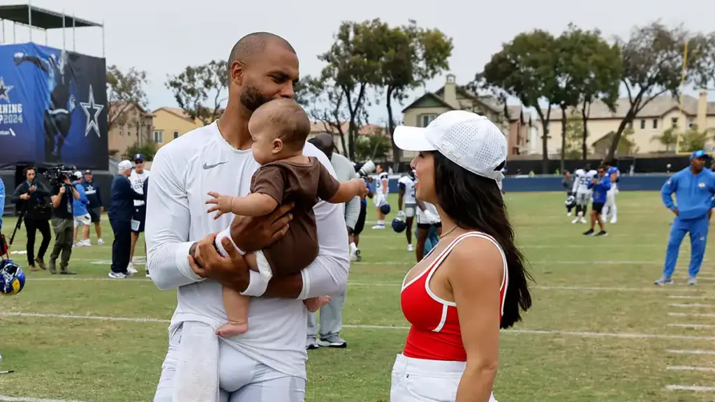 Referee in Dallas Cowboys uniform holding a baby and talking to a woman wearing a white cap and red sports top on the football field.