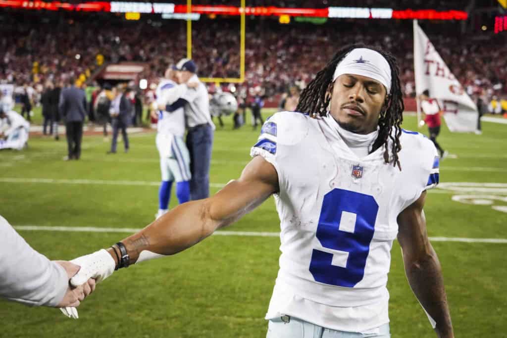 Helmetless Dallas Cowboys player on football field, holding hands with team member after game, showing sportsmanship and team unity, with cheering crowd in the background.