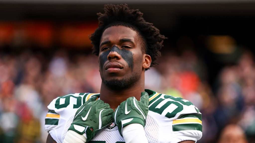 Young African American football player wearing green and white uniform with black eye paint, standing on the field, appearing focused, with a blurred crowd background at an NFL game.