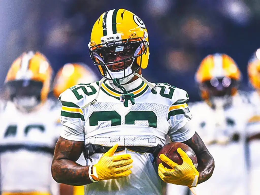 Green Bay Packers football player wearing helmet and uniform holding football, with teammates blurred in background, on game night.