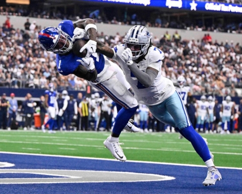 Catches football player in blue jersey being tackled by opponent in white jersey on football field during game.