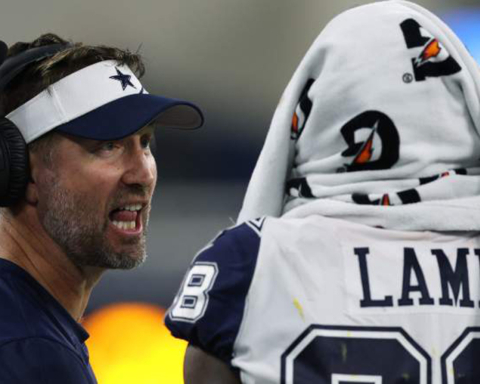 Dallas Cowboys coach with headset speaking to player wrapped in Gatorade towel at football game, inside AT&T Stadium.