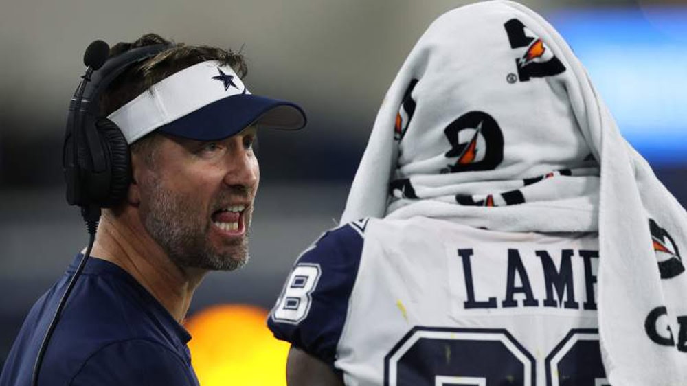 Dallas Cowboys coach with headset speaking to player wrapped in Gatorade towel at football game, inside AT&T Stadium.