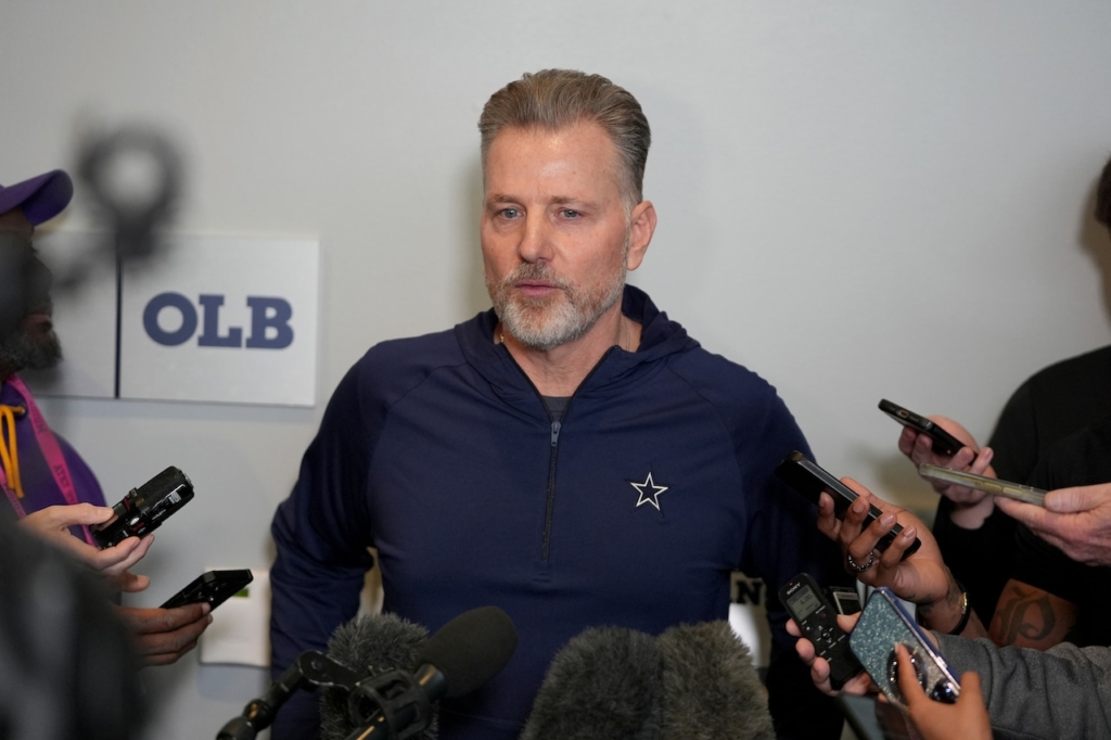 Dallas Cowboys coach speaking to the media during a press conference, surrounded by reporters with microphones and smartphones, inside the Inside The Star facility.