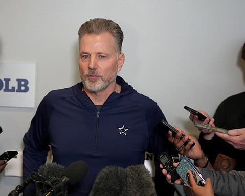 Dallas Cowboys coach speaking to the media during a press conference, surrounded by reporters with microphones and smartphones, inside the Inside The Star facility.