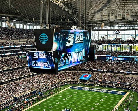 Bright indoor football stadium with large digital scoreboard displaying "3rd Down", surrounded by a packed crowd at AT&T Stadium Dallas Cowboys.