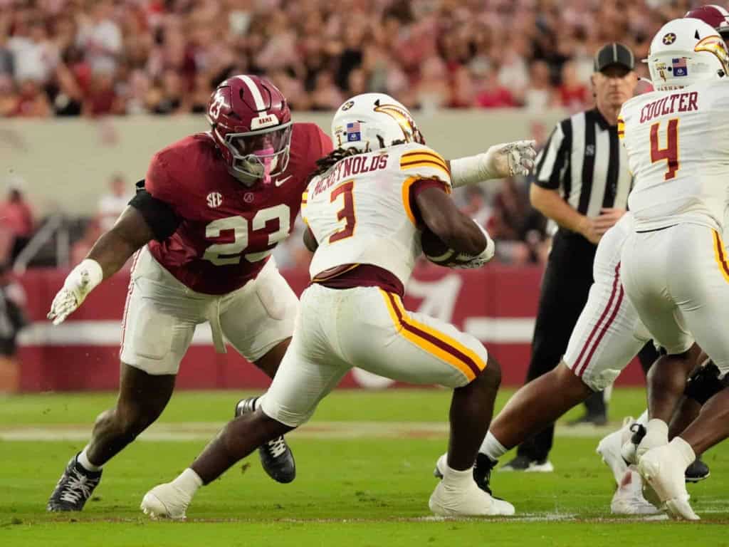 Alabama football linebacker #23 tackling a Kansas City Chiefs running back #3 during a game at a packed stadium.