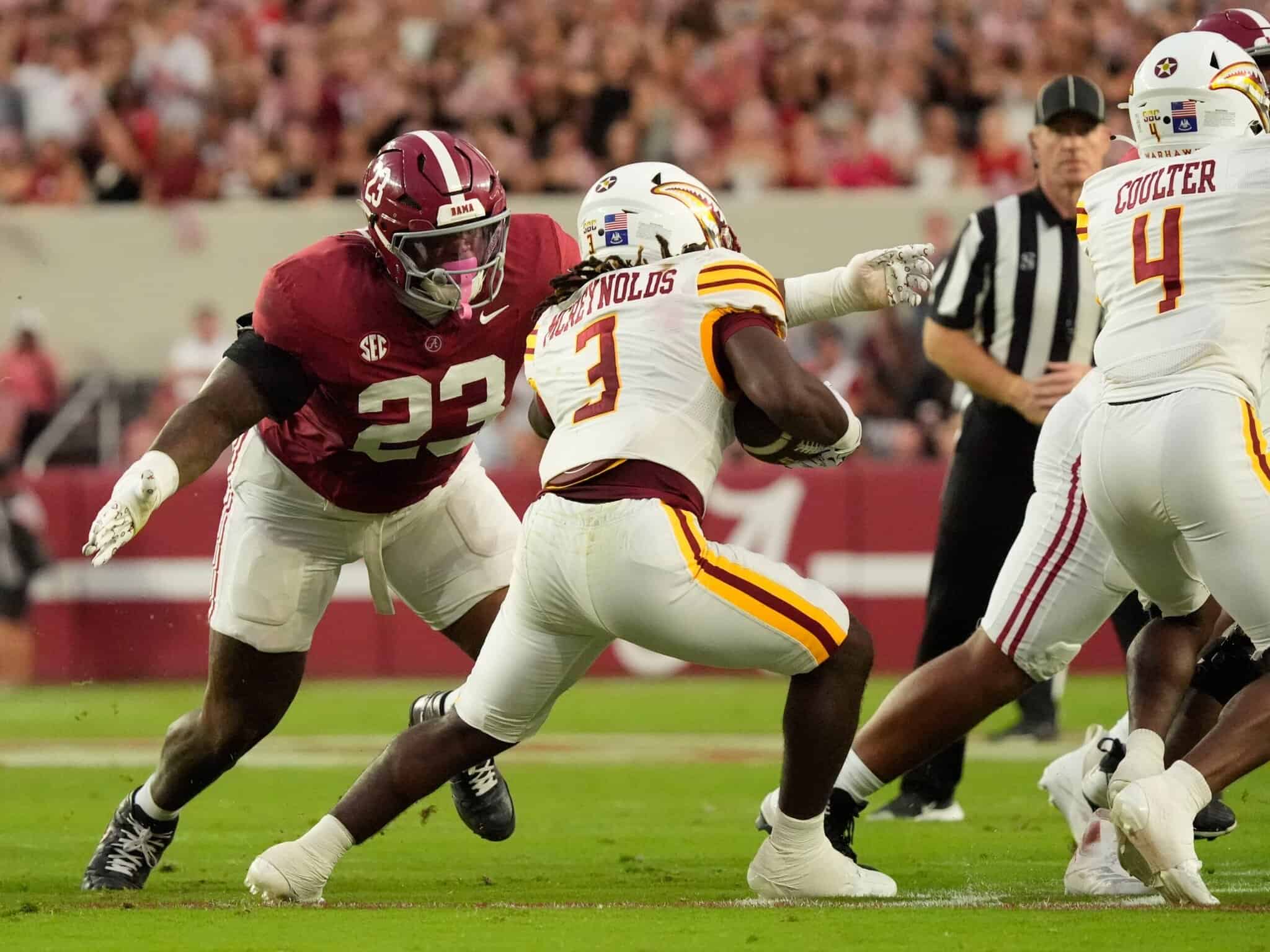 Alabama football linebacker #23 tackling a Kansas City Chiefs running back #3 during a game at a packed stadium.