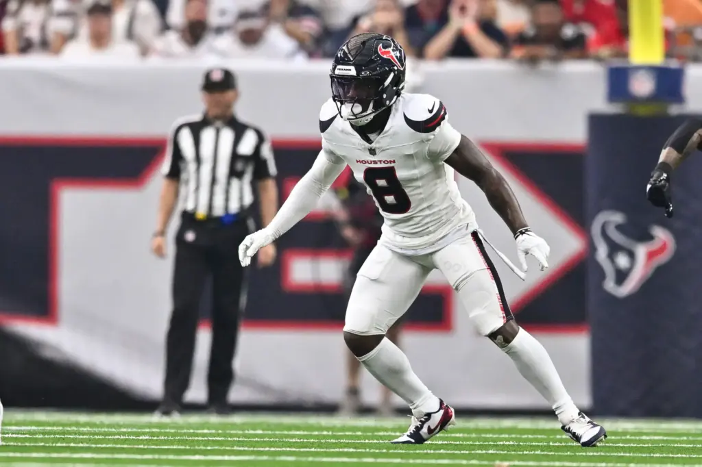 Houston Texans football player during game action, wearing white jersey number 8, on the field at NRG Stadium in Houston, Texas, with a blurred crowd and referee in the background.