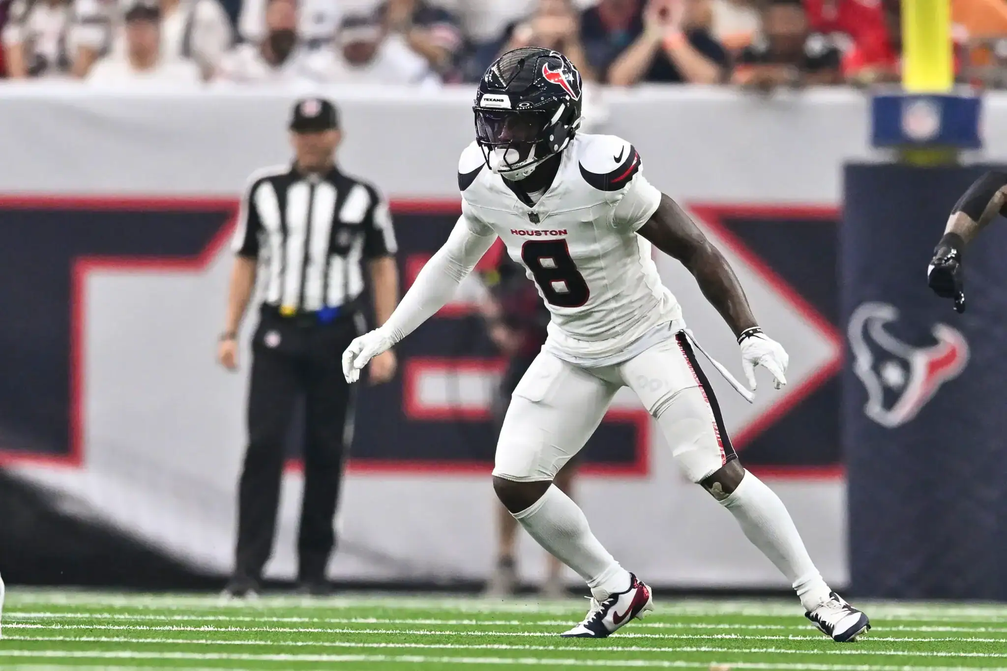 Houston Texans football player during game action, wearing white jersey number 8, on the field at NRG Stadium in Houston, Texas, with a blurred crowd and referee in the background.