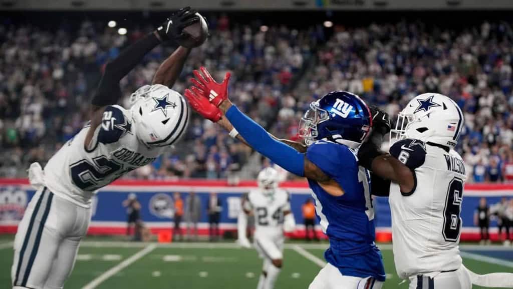 Dallas Cowboys football player catching a pass against New York Giants defenders during an NFL game. Action shot showing intense gameplay at the stadium with fans in the background.