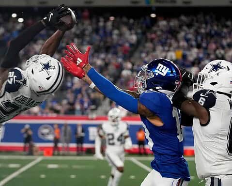 Dallas Cowboys football player catching a pass against New York Giants defenders during an NFL game. Action shot showing intense gameplay at the stadium with fans in the background.