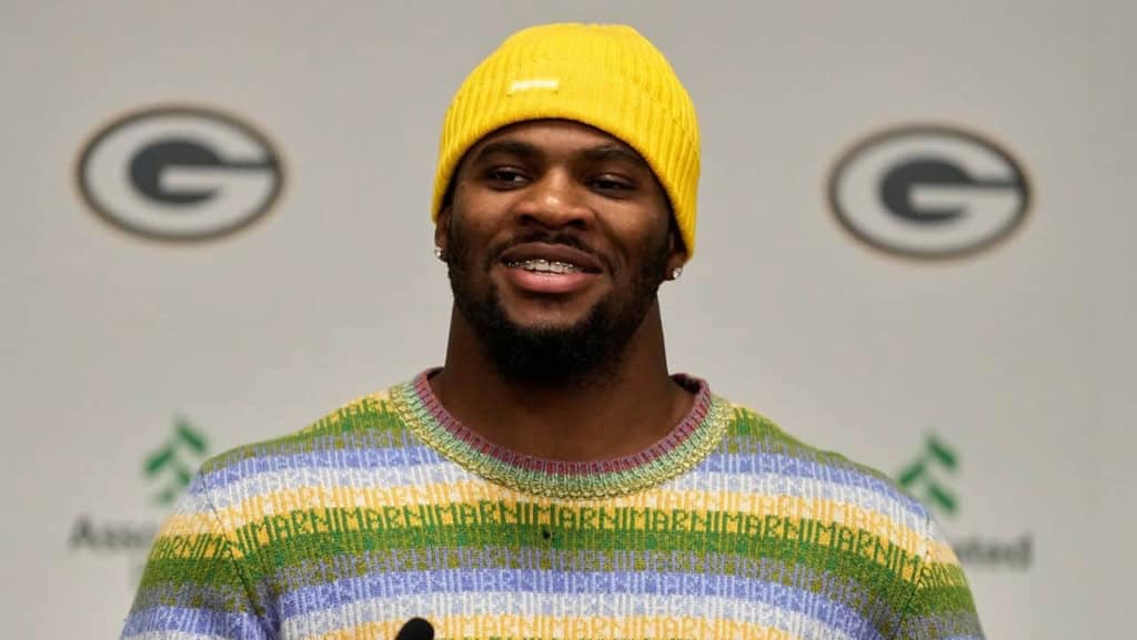 A man wearing a bright yellow beanie and colorful sweater speaking at a press conference with the Green Bay Packers logo in the background.