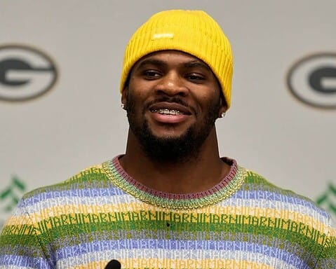 A man wearing a bright yellow beanie and colorful sweater speaking at a press conference with the Green Bay Packers logo in the background.