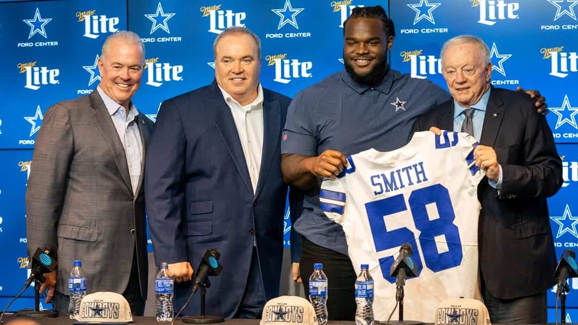 Dallas Cowboys player holding a jersey at a press conference with executives, inside the Ford Center at The Star, promoting NFL team news and Dallas Cowboys updates.