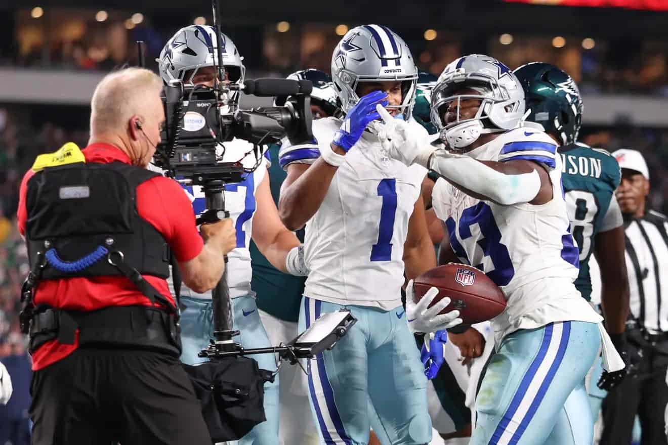 Celebrating a touchdown at an NFL game, Dallas Cowboys players cheer as a teammate scores, with a cameraman capturing the victorious moment during the football game.