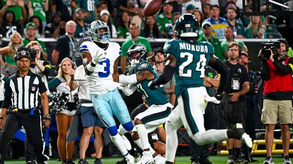 Bright football action featuring Dallas Cowboys and Philadelphia Eagles players during a game, with cheerleaders, referees, and spectators in the background at a packed stadium.