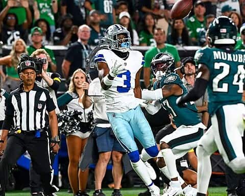 Bright football action featuring Dallas Cowboys and Philadelphia Eagles players during a game, with cheerleaders, referees, and spectators in the background at a packed stadium.