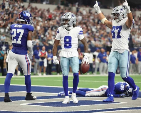 The Dallas Cowboys celebrate a touchdown on the field during an NFL game against the New York Giants. The players are wearing their team uniforms and helmets, with a packed stadium crowd in the background. The scene captures the excitement and intensity of professional football.