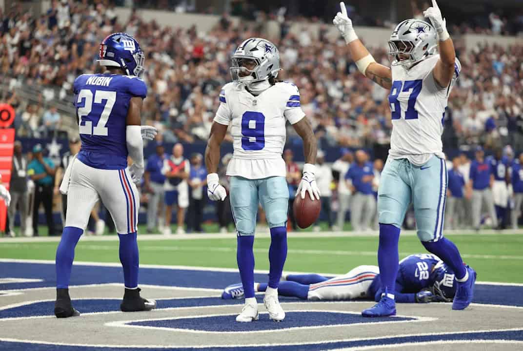 The Dallas Cowboys celebrate a touchdown on the field during an NFL game against the New York Giants. The players are wearing their team uniforms and helmets, with a packed stadium crowd in the background. The scene captures the excitement and intensity of professional football.