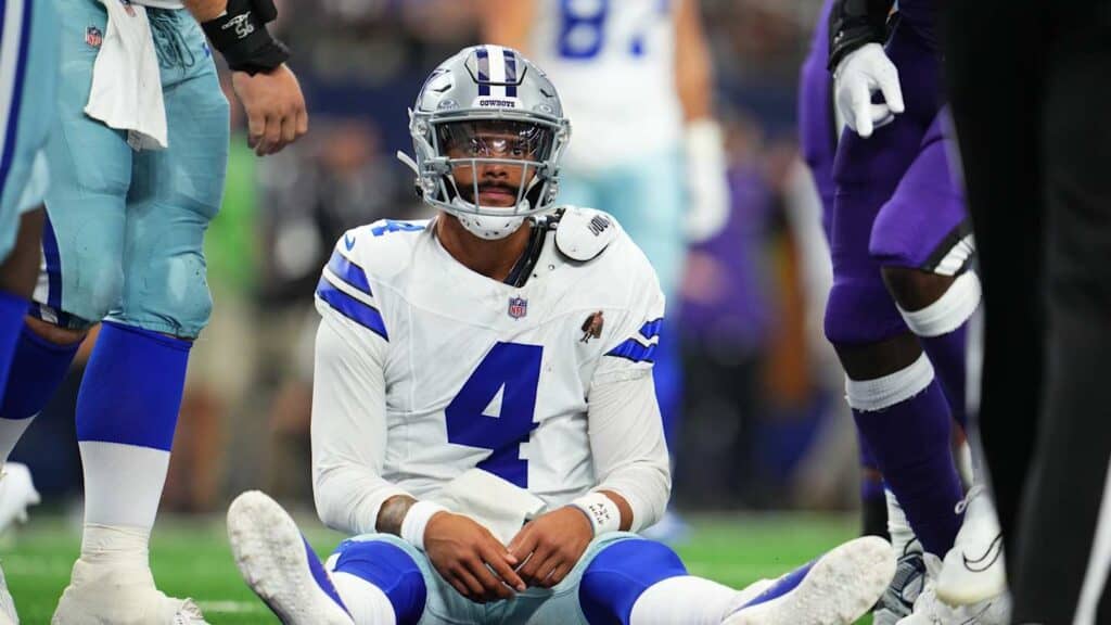 Dallas Cowboys player wearing jersey number 4 sitting on the field with teammates standing around him after injury.