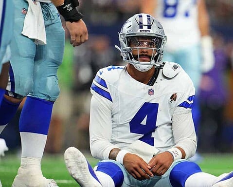 Dallas Cowboys player wearing jersey number 4 sitting on the field with teammates standing around him after injury.