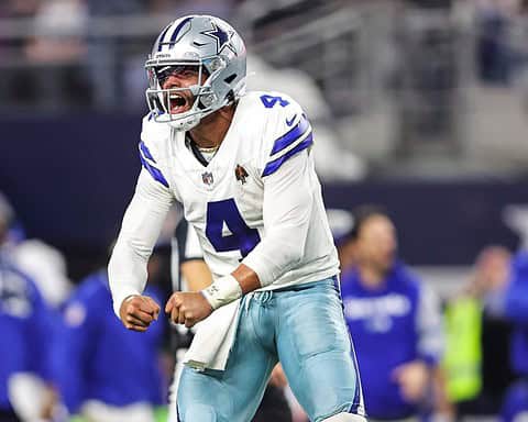 Celebrating Dallas Cowboys player player Derek Carr during a game, showing intense emotion and excitement, wearing team jersey and helmet, with teammates and fans in the background.