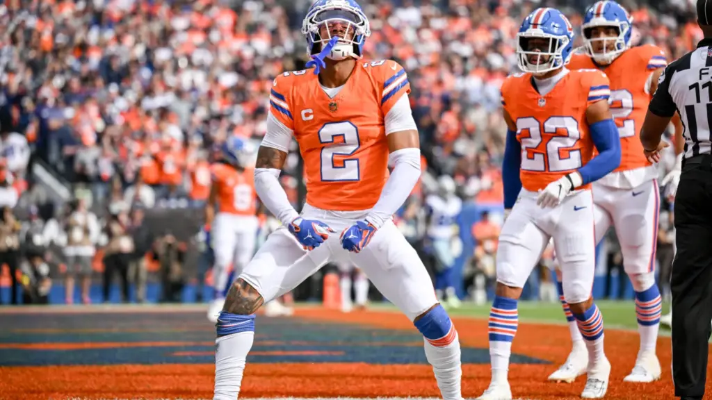 Celebrating a touchdown on the field, Denver Broncos players in their orange and white uniforms during an NFL game. The crowd cheers loudly in the background at Empower Field at Mile High.