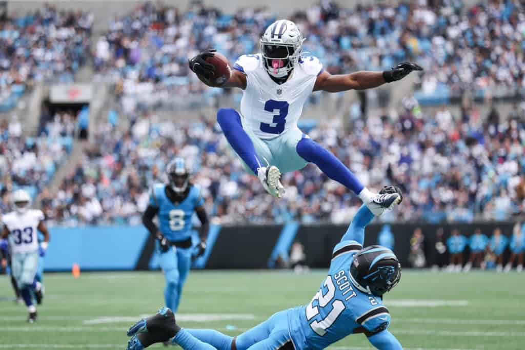 High-action Dallas Cowboys football player jumping over opposing Carolina Panthers player during a game at AT&T Stadium, showcasing athleticism and team rivalry.