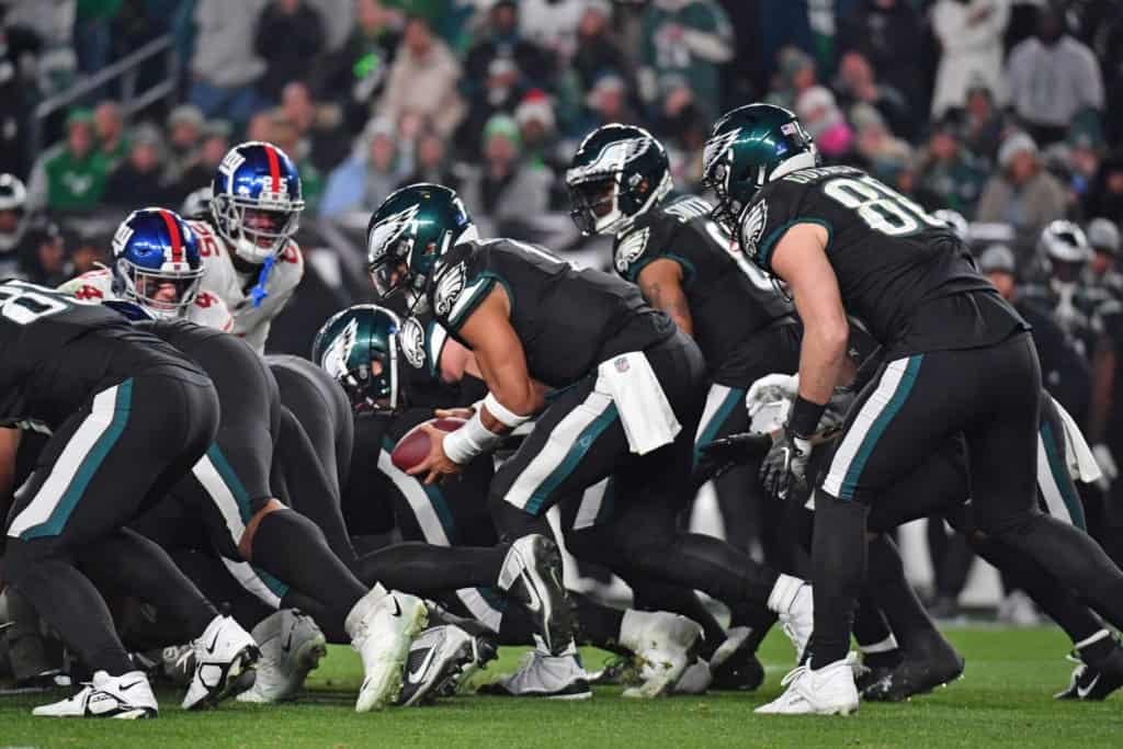 Playing American football at Lincoln Financial Field, featuring the Philadelphia Eagles' offensive line and the opposing defense, with a packed stadium in the background.