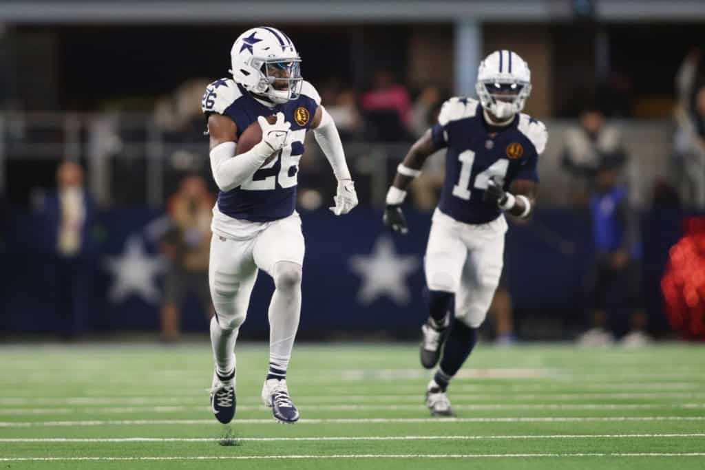Running back Dallas Cowboys football player wearing jersey number 22, in an action shot during a game on the field, holding a football, with teammate number 14, in a stadium setting.