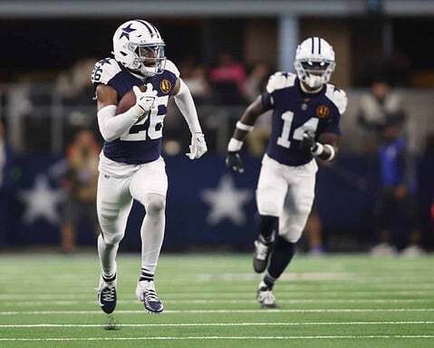 Running back Dallas Cowboys football player wearing jersey number 22, in an action shot during a game on the field, holding a football, with teammate number 14, in a stadium setting.
