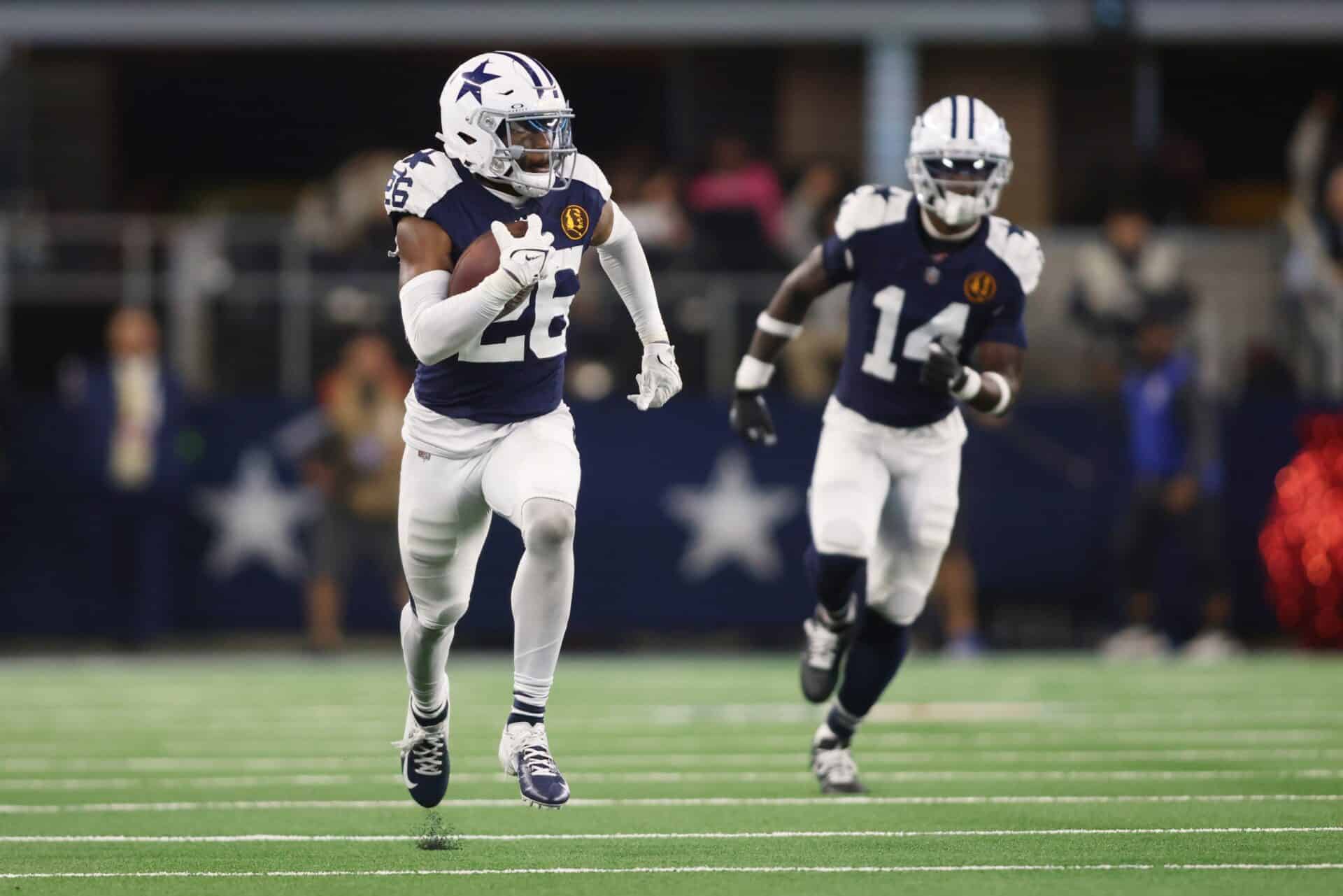Running back Dallas Cowboys football player wearing jersey number 22, in an action shot during a game on the field, holding a football, with teammate number 14, in a stadium setting.
