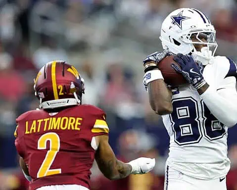 Outside linebacker leading Dallas Cowboys onto the field during a game, holding football, wearing team gear, with stadium crowd in background.