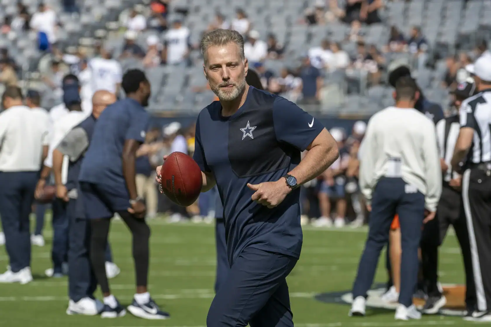 Outside linebacker leading Dallas Cowboys onto the field during a game, holding football, wearing team gear, with stadium crowd in background.