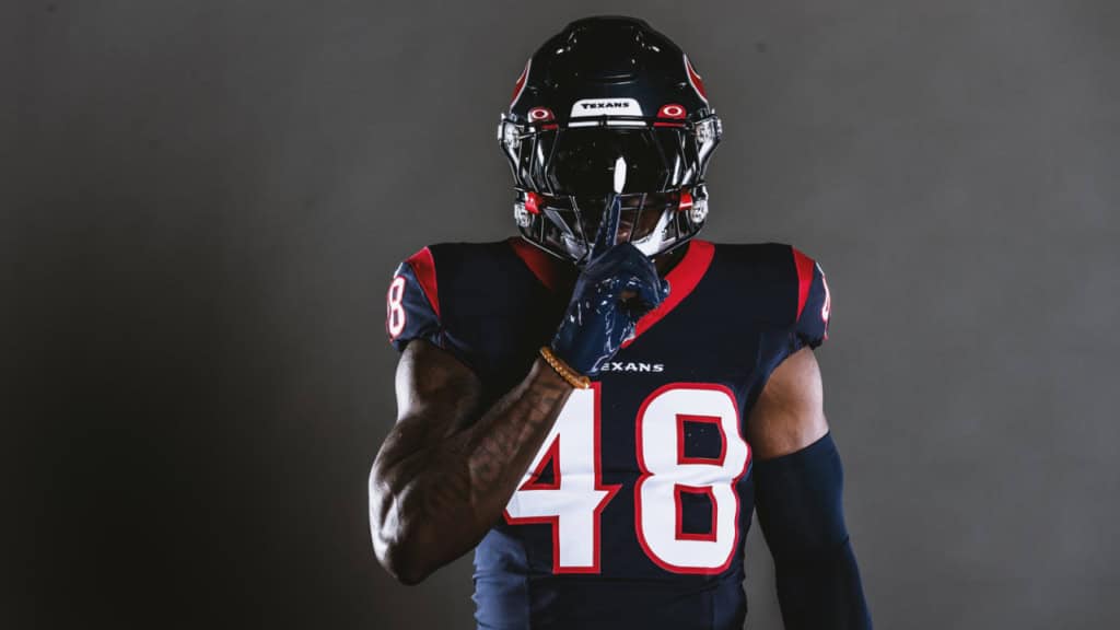 Young Houston Texans football player wearing jersey number 48, standing in a dark studio setting, posing with finger to lips, showcasing team spirit and athletic strength.