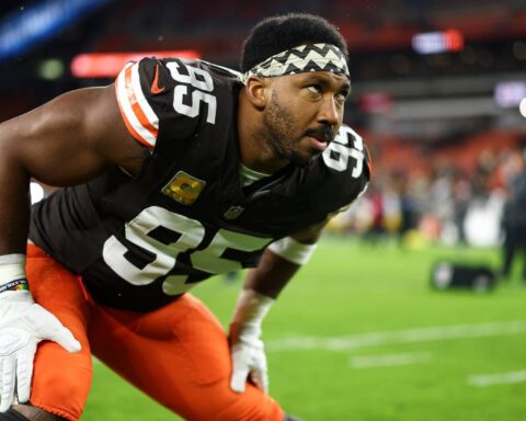 Athlete in football uniform crouching on the field, wearing number 55 for the Dallas Cowboys, preparing for the game with a focused expression, stadium lights, and fans in the background.