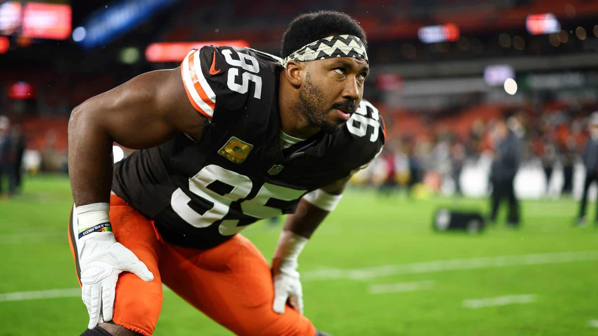 Athlete in football uniform crouching on the field, wearing number 55 for the Dallas Cowboys, preparing for the game with a focused expression, stadium lights, and fans in the background.